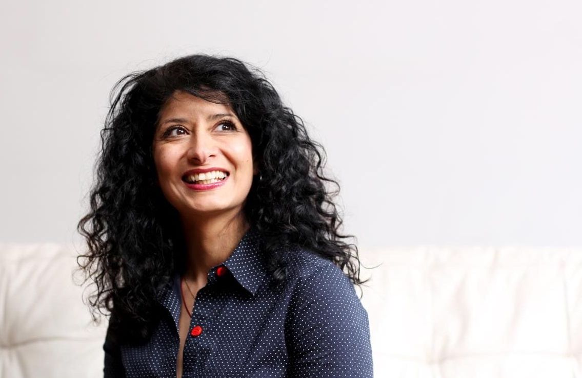 Shappi Khorsandi smiling and looking at a distance. She is sitting on a large couch wearing a buttoned up shirt and has wavy curly hair.