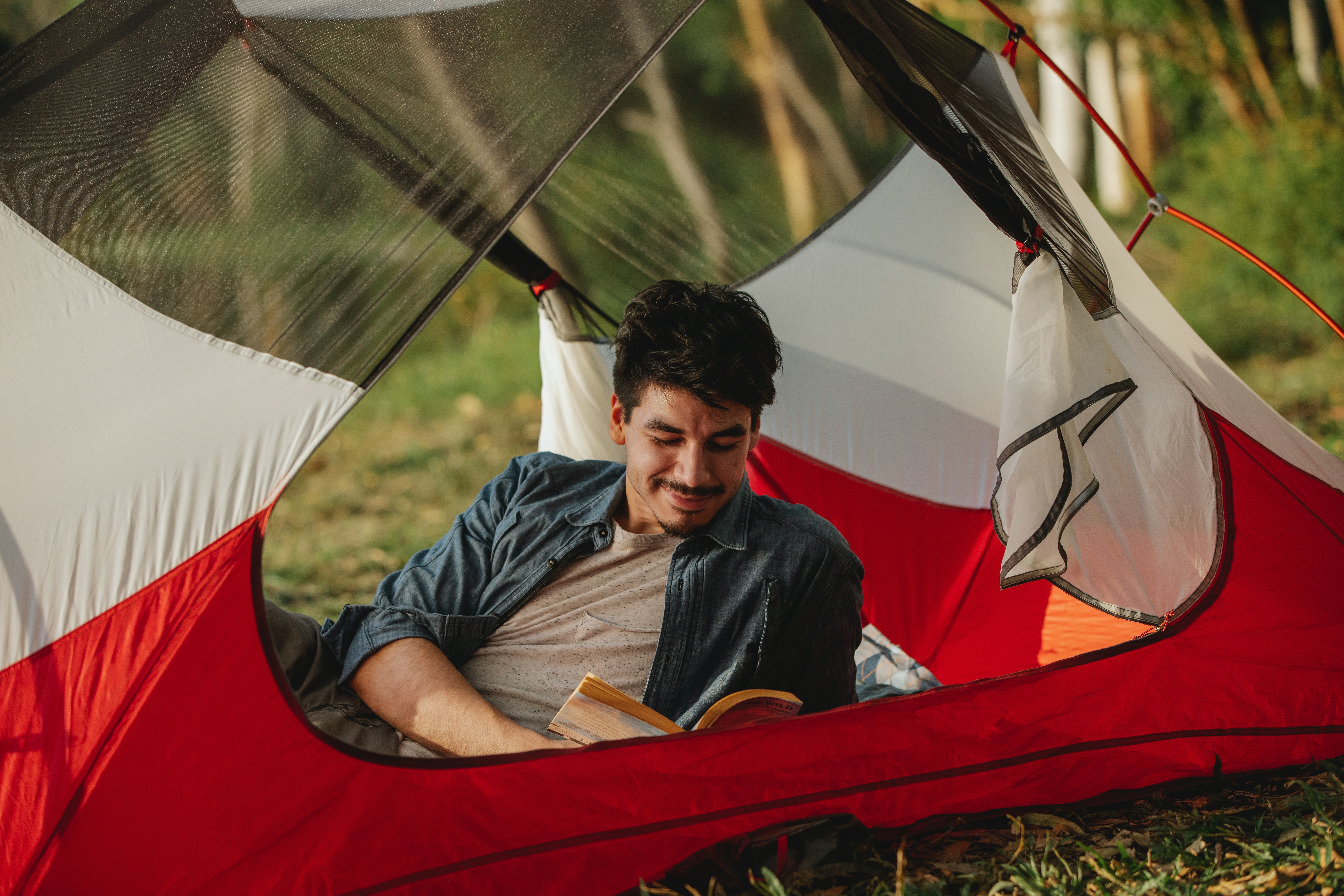 A young man smiles while reading a book inside his tent in the outdoors.