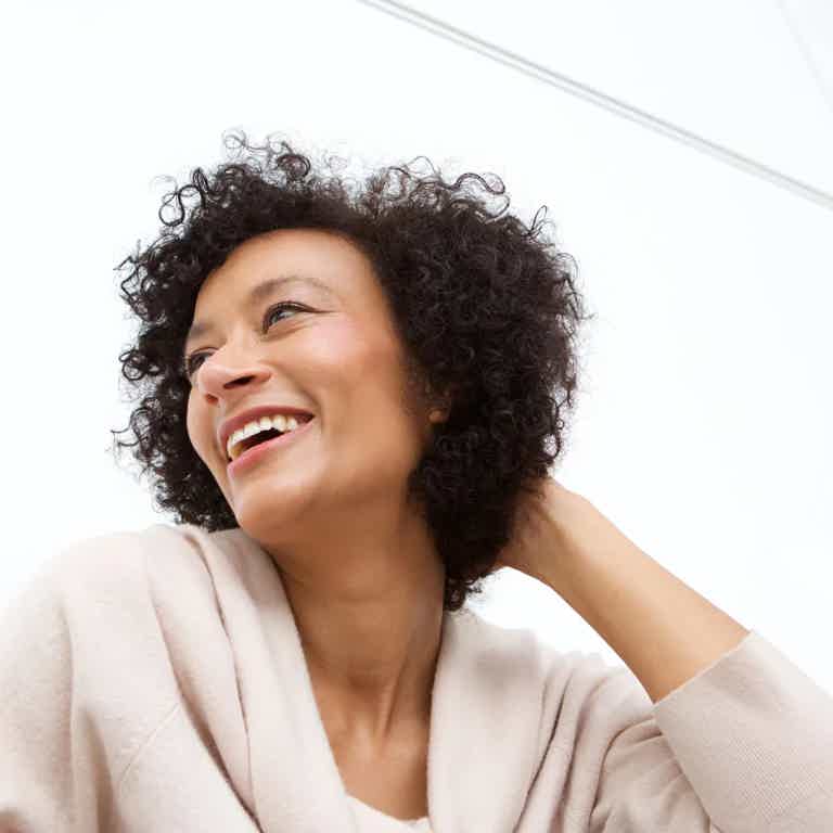 Woman with curly hair smiling, looking to the side, wearing a light sweater indoors.