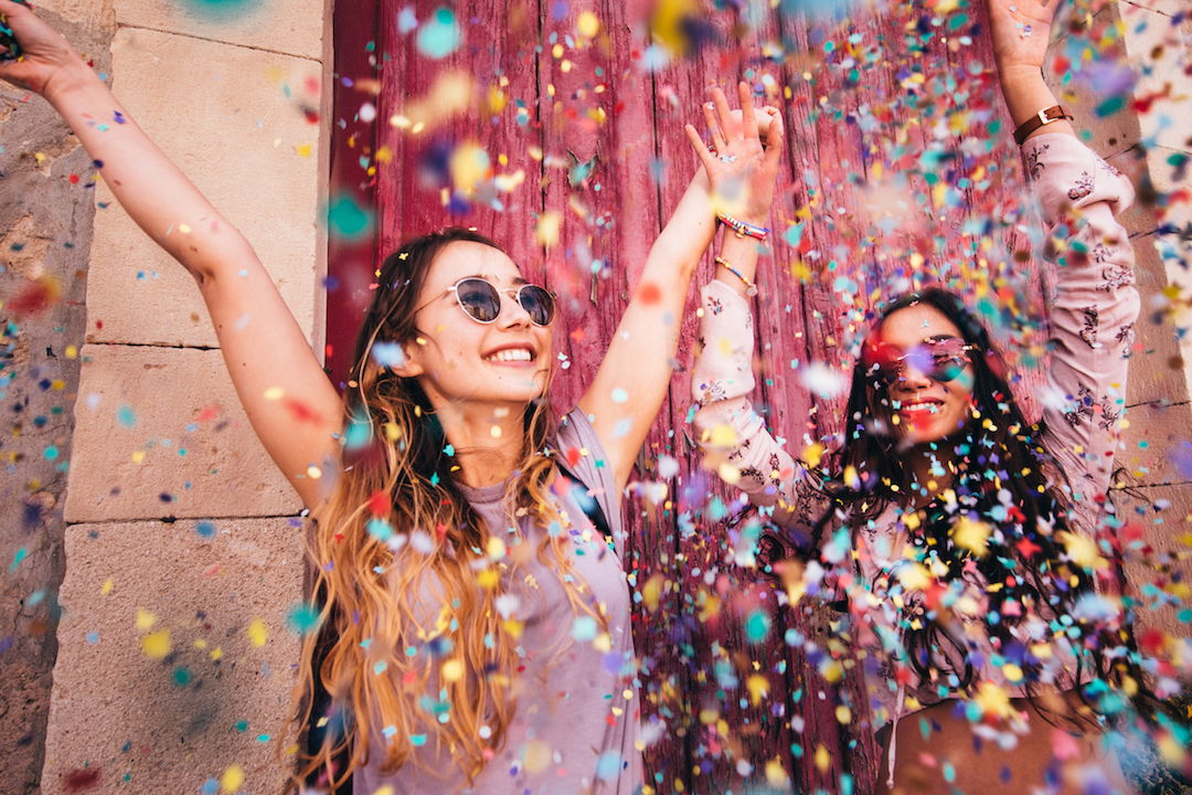 Two young women with sunglasses with their hands in the air and a lot of colorful paper and glitter flying everywhere.