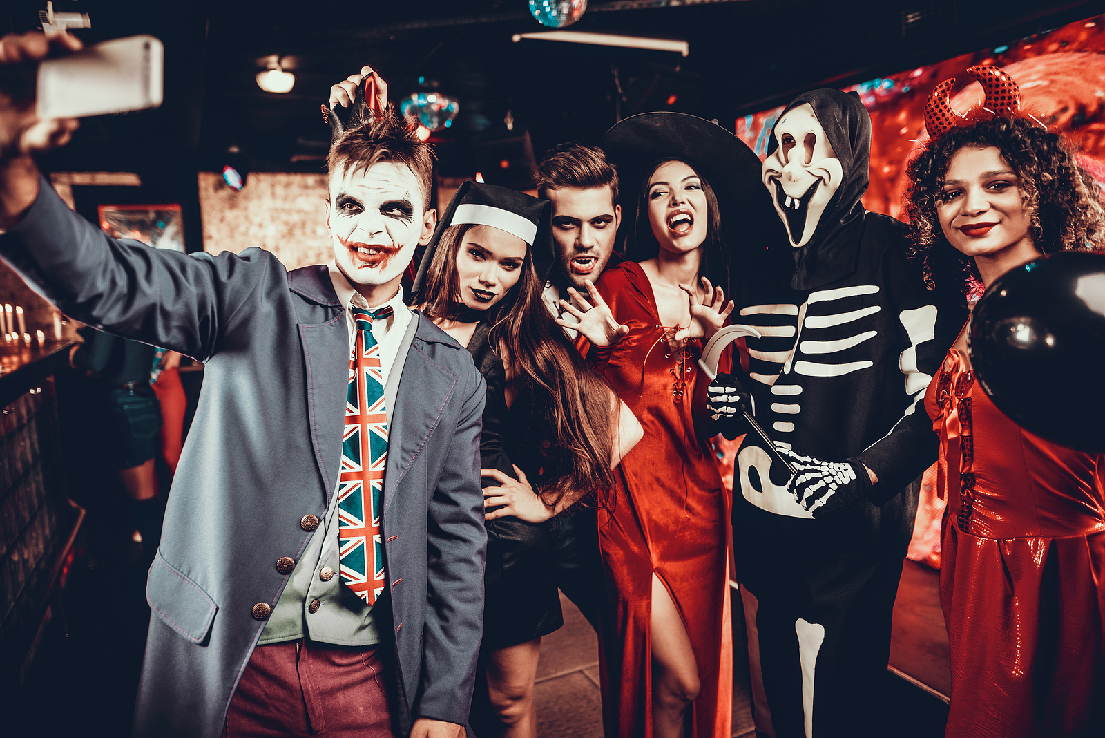 A young man takes a selfie with his friends who are in costume at a Halloween party at a bar.