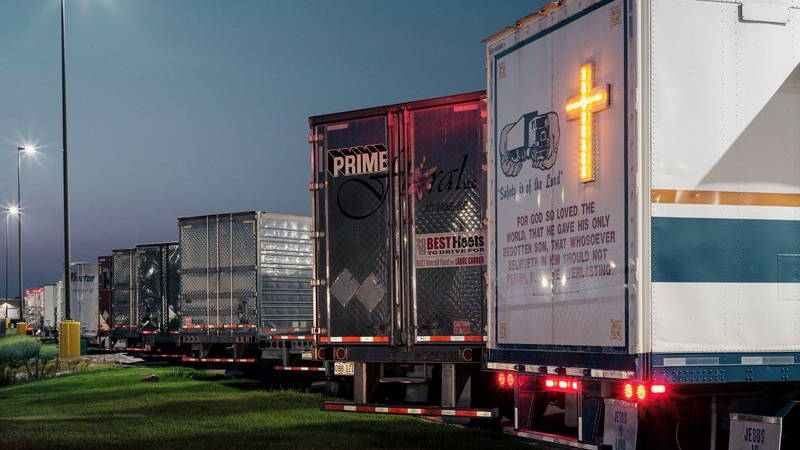 Line of semi-trucks in a parking lot shot from behind; one truck, the Mobile Chapel, has a lit-up cross and bible verse on the back