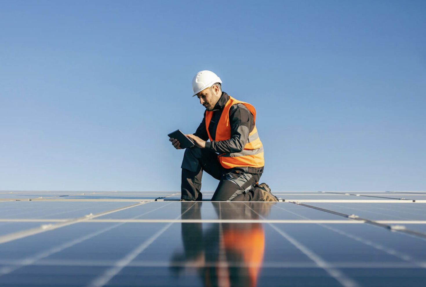 A person wearing a white hard hat and orange safety vest kneels on solar panels while using a tablet device, with a clear blue sky in the background.
