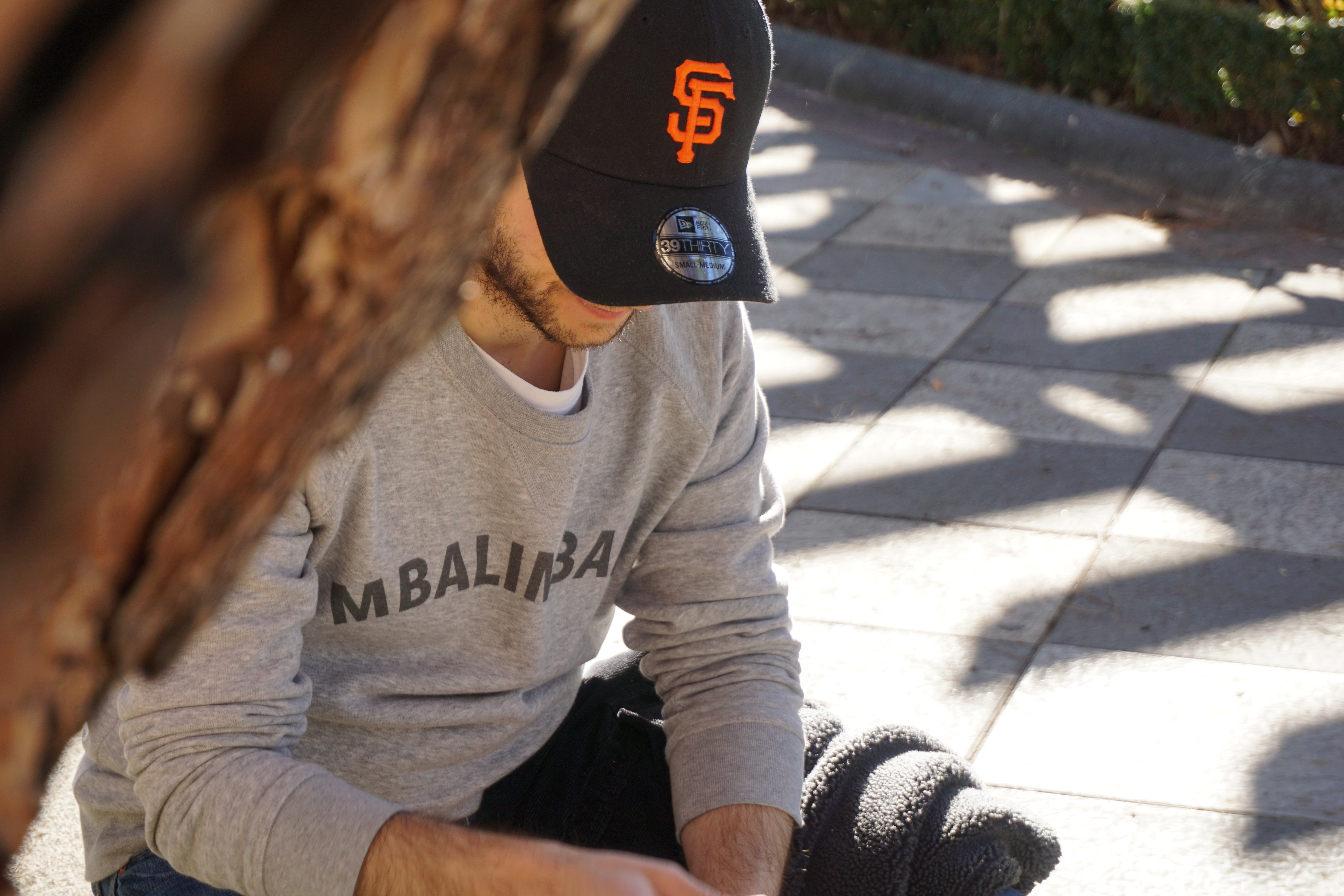 A man sits down at a bench outdoors looking down wearing a baseball cap with his face half covered.