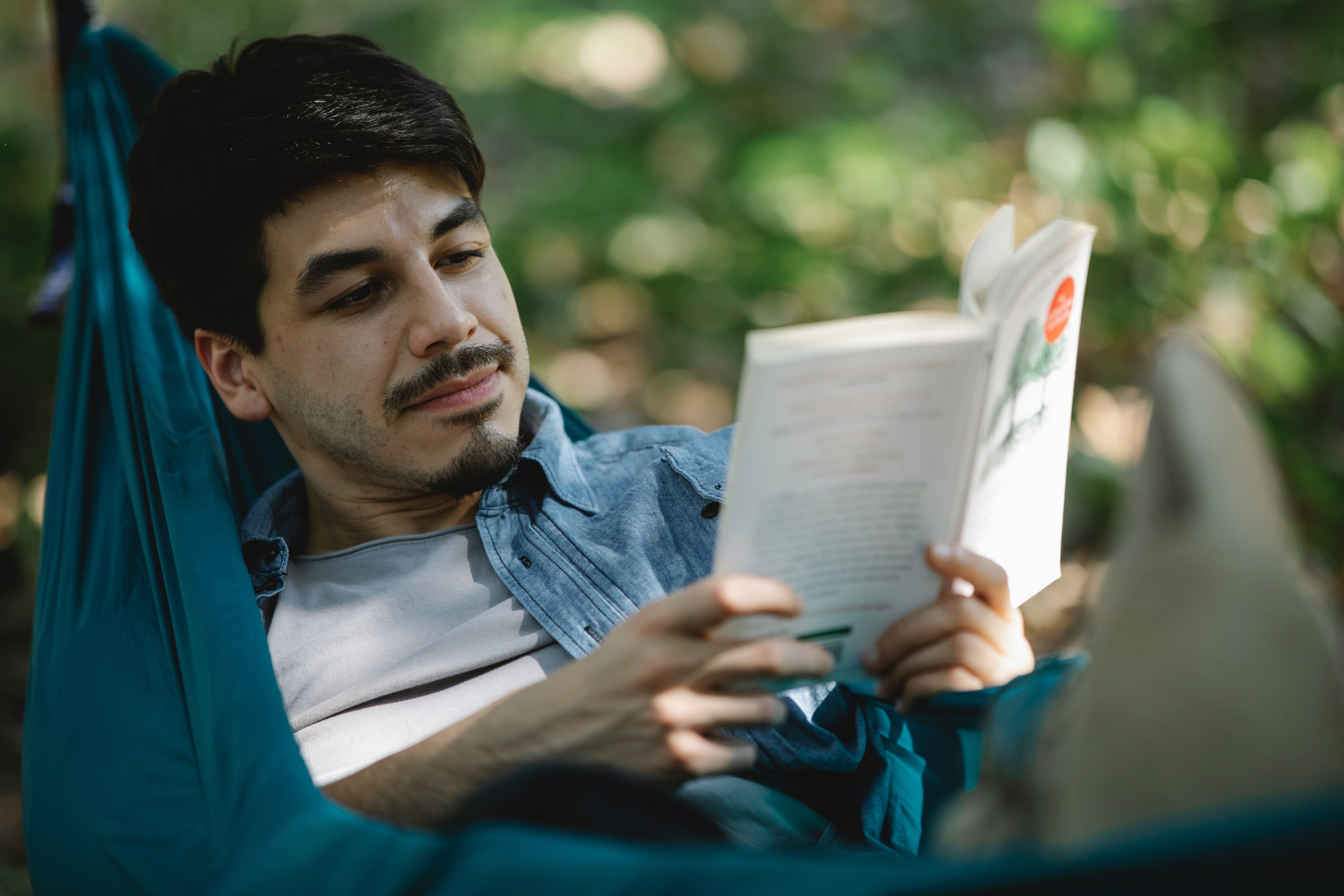 A multi ethnic man lays on a hammock outside and reads a book smiling.