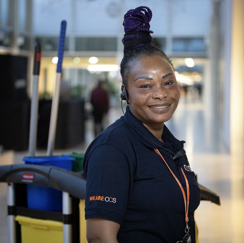 A smiling woman in a cleaning uniform stands in a bright hallway near cleaning equipment, with brooms and a mop visible behind her. She wears a headset and a lanyard, and her hair is styled in purple braids.