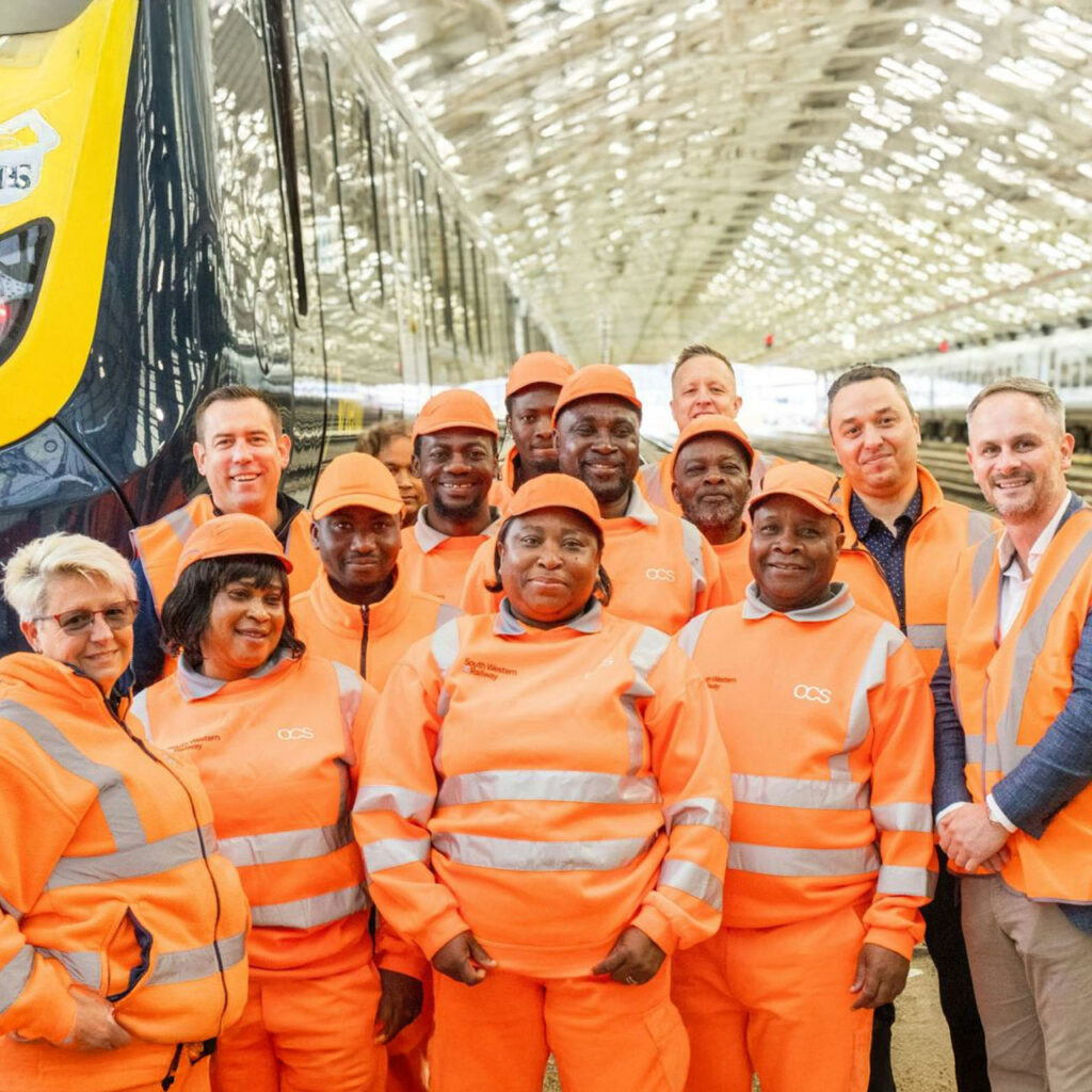 A group of rail workers in orange high-visibility uniforms and hard hats pose together, smiling, inside a train station next to a train.