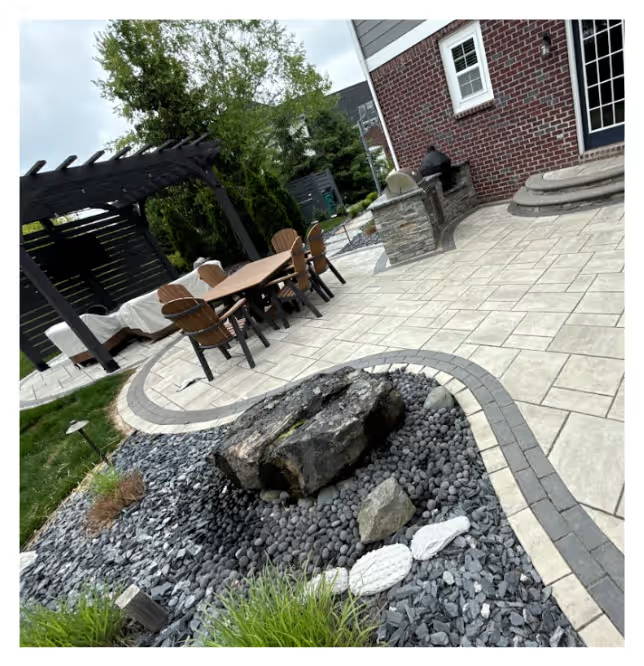 Modern backyard patio with large stone pavers, curved border detailing, central rock feature with decorative gravel, outdoor dining set under a pergola, and built-in grill beside a brick house.