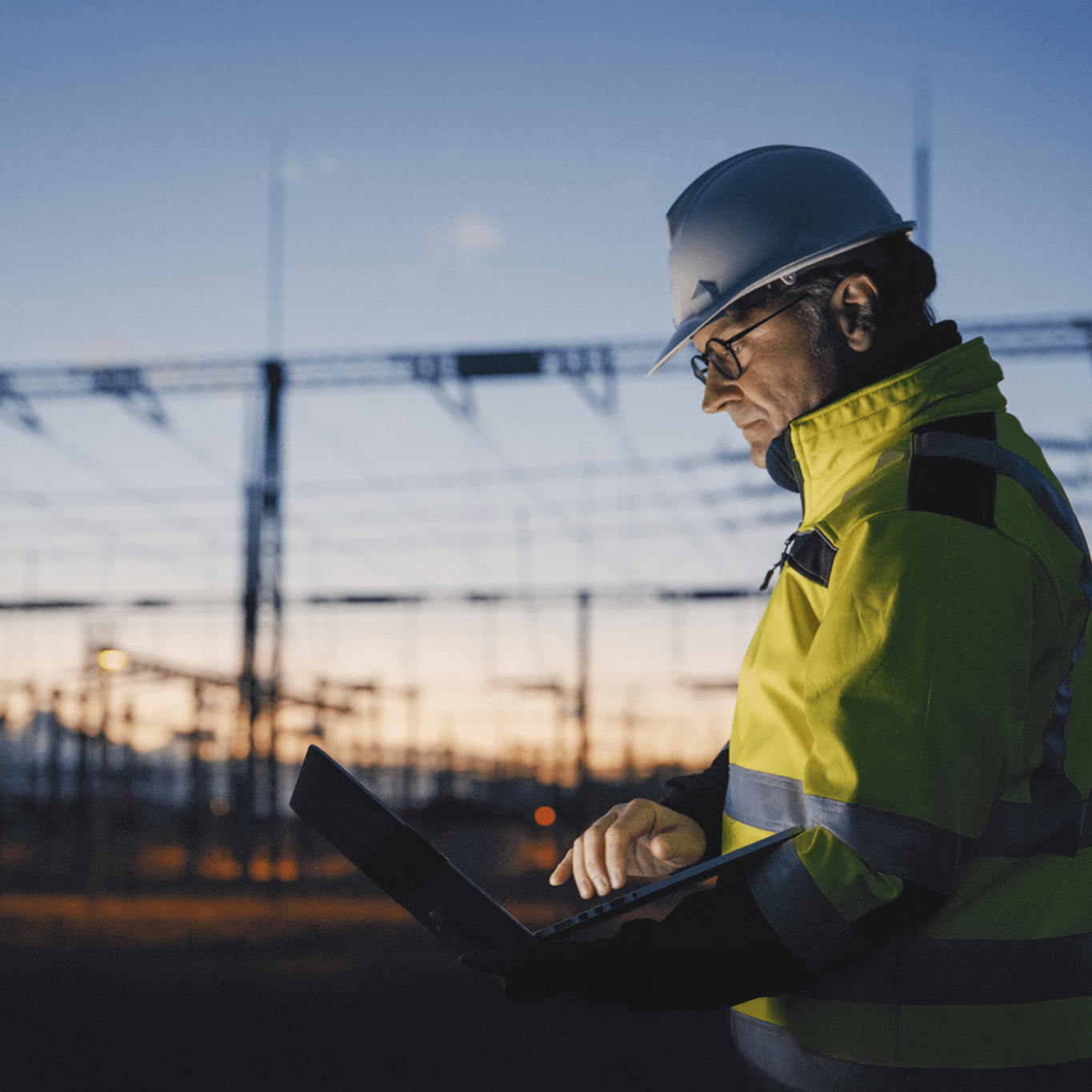 A worker in a high-visibility jacket and hard hat uses a laptop outdoors at dusk, with power lines and electrical structures in the background.