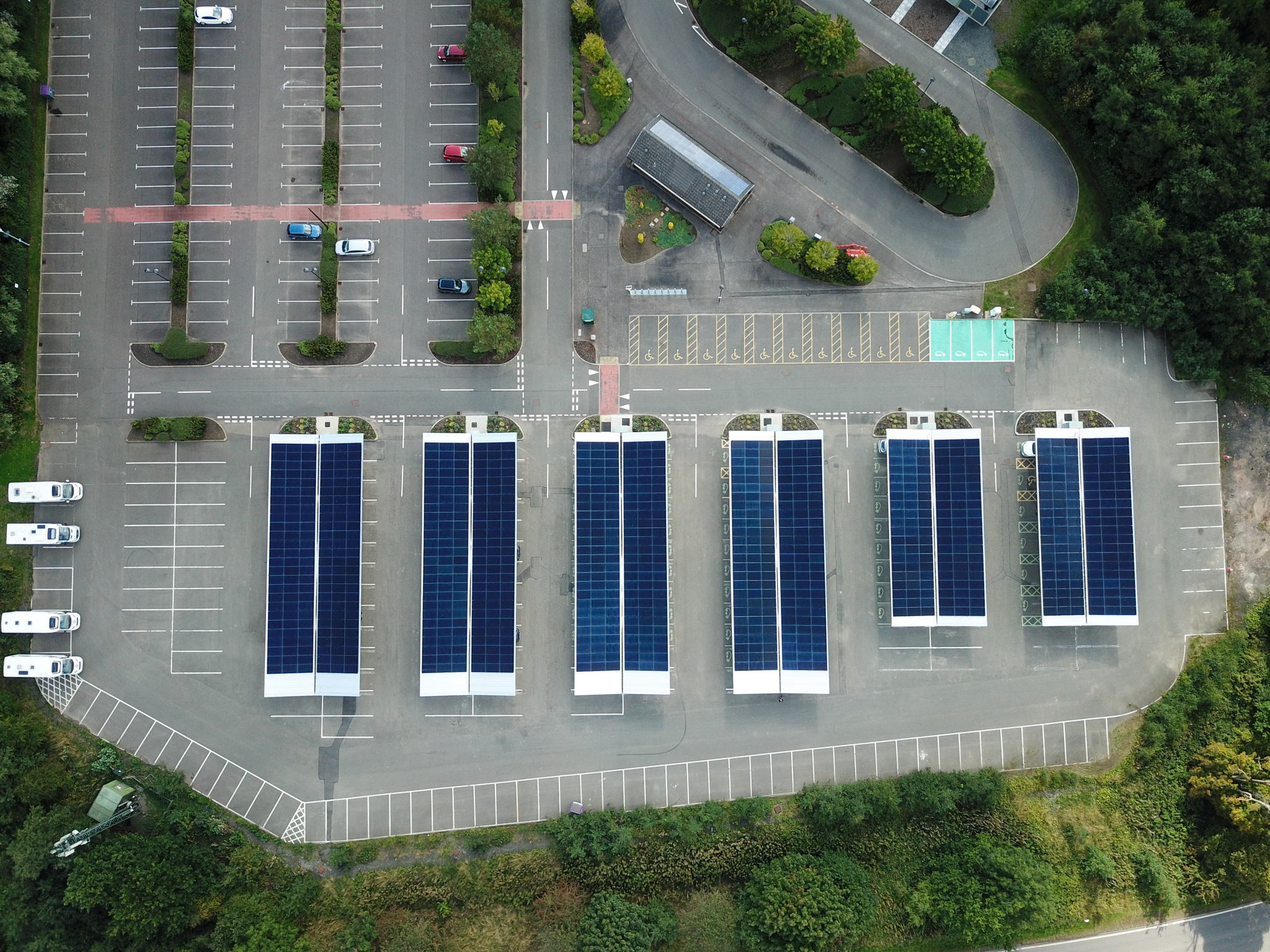 Aerial view of a parking lot with several large rows of solar panel canopies, surrounded by trees, with most parking spaces unoccupied.