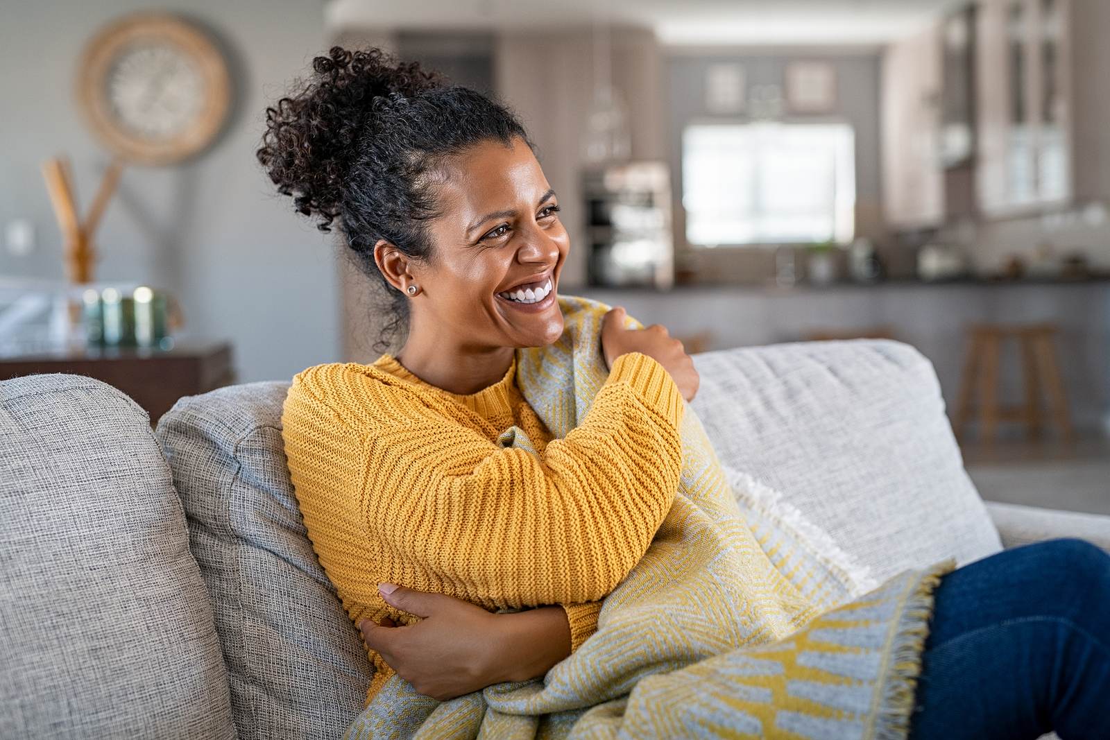A black woman sits on her couch watching tv and laughing hugging a pillow.