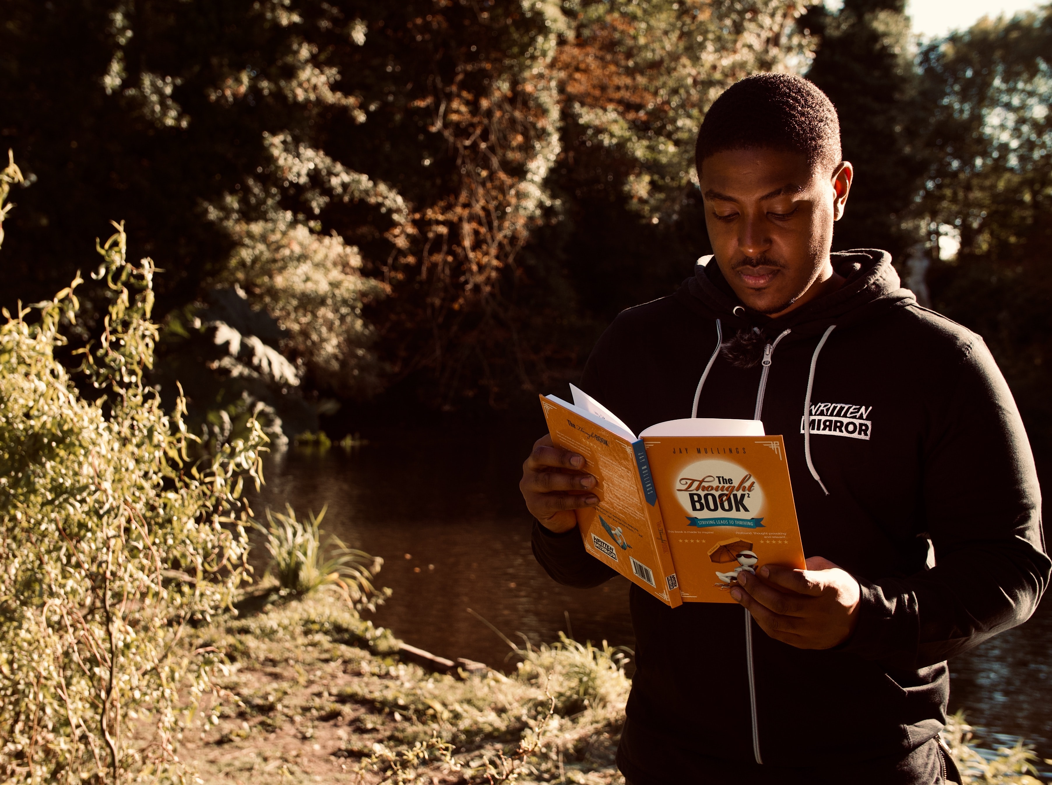 A black man walks in the outdoors while reading a book.