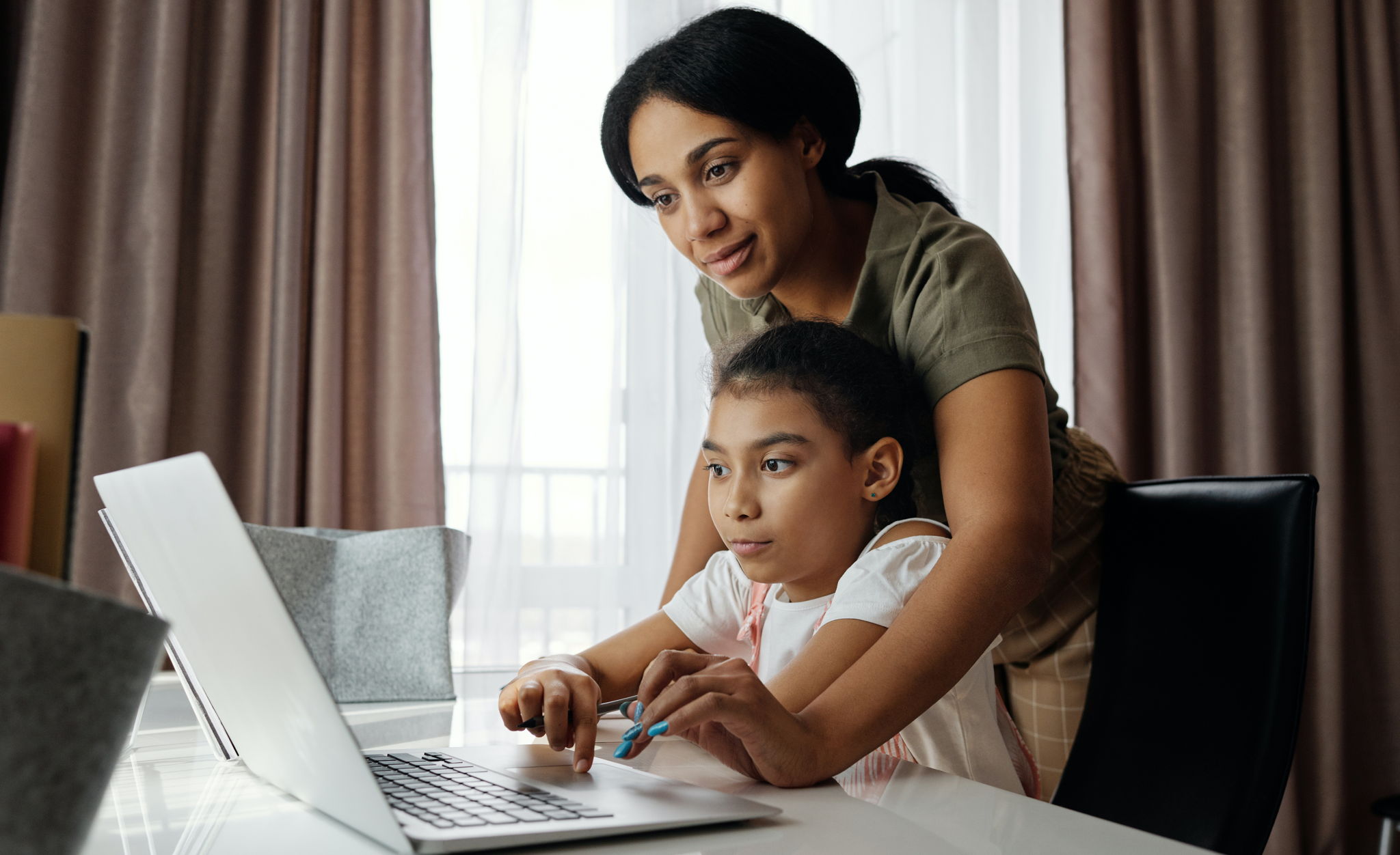 A multi ethnic mom and her daughter, looking at a laptop together at a desk. The mom is guiding the child showing her things on the computer.