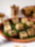 Squares of Indian barfi garnished with crushed pistachios on a banana leaf–lined clay plate, with a brass lamp and cup in the background.
