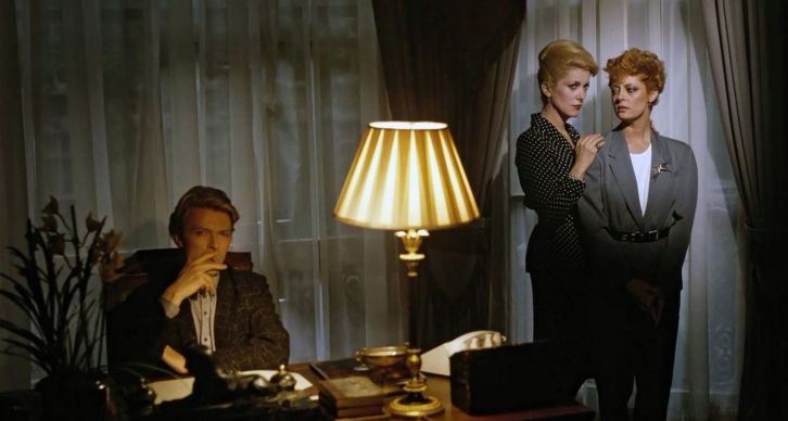 Catherine and Susan standing to the side together looking over at Bowie who is sitting at his desk hand on his mouth.