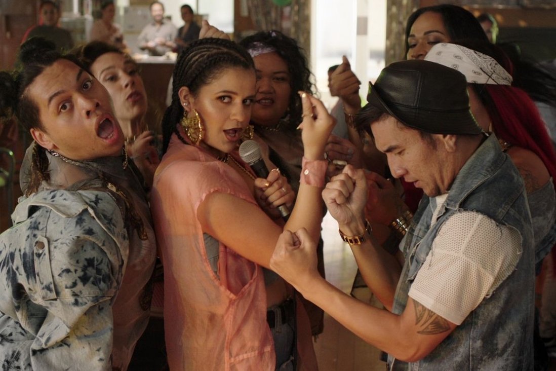 Image of several of the cast members who are POC, looking at the camera while they are dancing together during the day inside a building. Everyone has on fashionable clothes.
