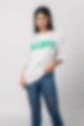 A woman wearing a white T-shirt with green ‘TALENTED’ lettering, paired with jeans, on a neutral background, showcasing a casual style.