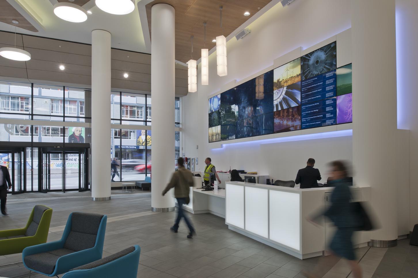 Modern office lobby with people walking and standing near a white reception desk; large digital display screens on the wall show various images and information; floor-to-ceiling windows provide natural light.