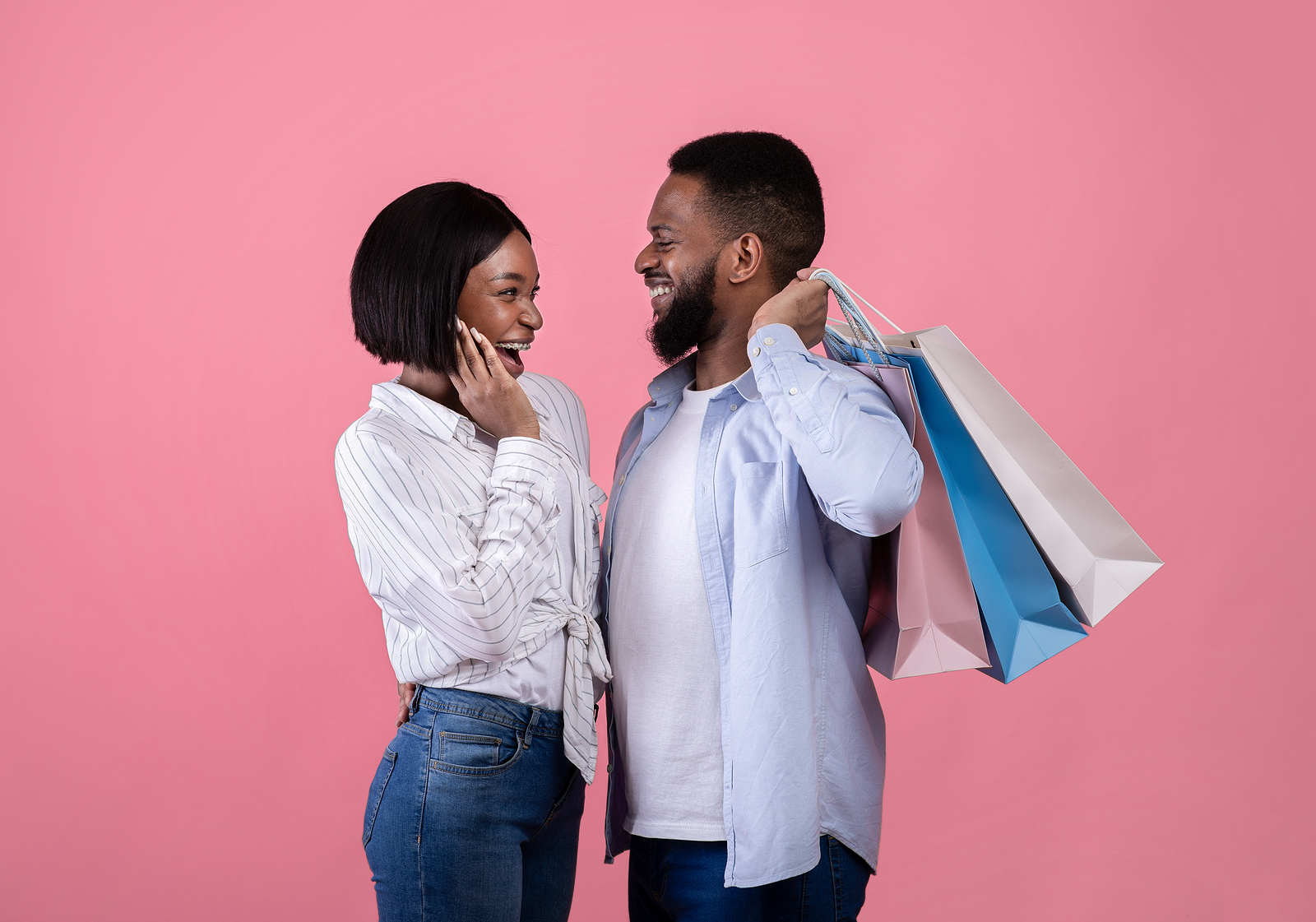 A young black couple smiling facing eachother. She has a surprised expression while he smiles holding shopping bags behind his back.