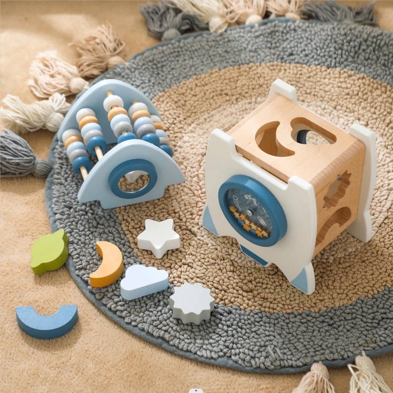 Une collection de fusées d'apprentissage Montessori en bois InvenToy, comprenant un labyrinthe de perles, un trieur de formes et des blocs à empiler, disposés sur un tapis tissé circulaire. Les jouets présentent une palette de couleurs neutres avec des touches de bleu.