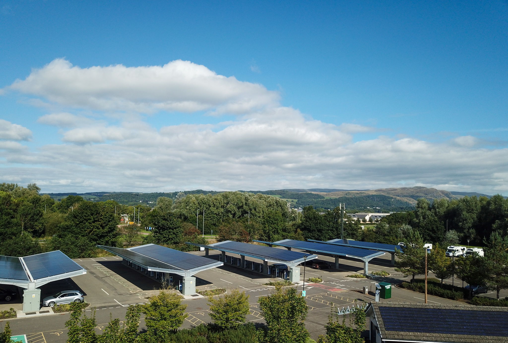 A parking lot with several carports covered in solar panels, surrounded by trees and greenery, under a partly cloudy blue sky with hills visible in the background.