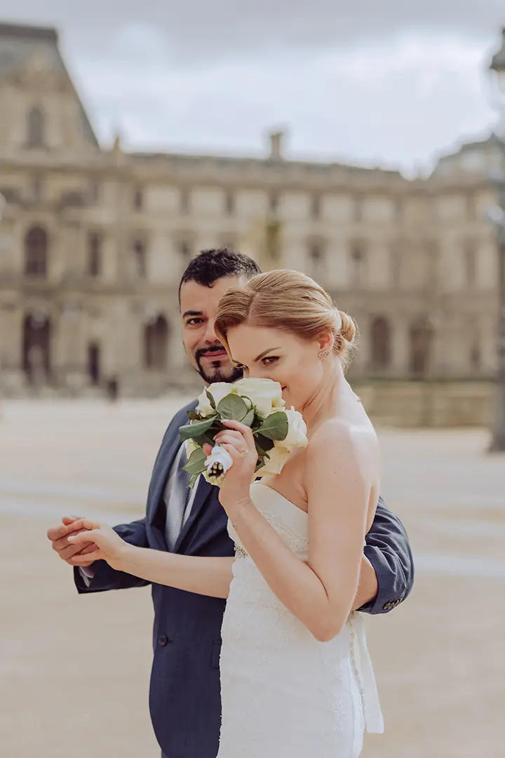 La mariée tenant un bouquet et un marié posent devant le Louvre pour la photo.