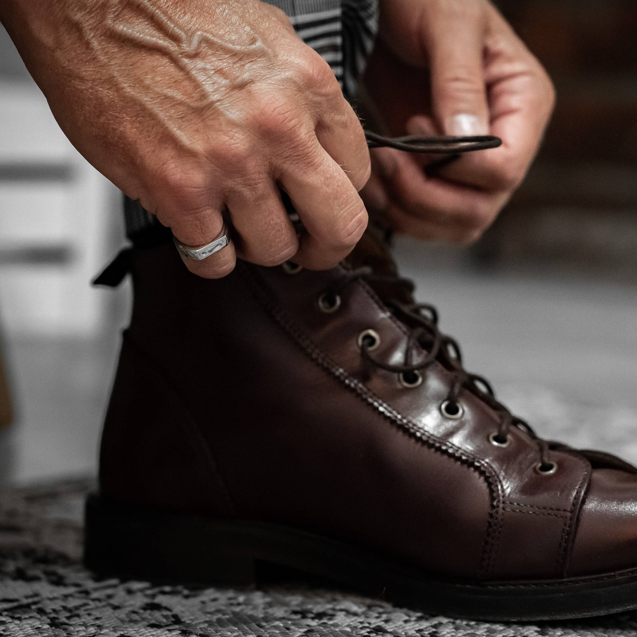Close up image of a brown lace up boot and a man's hands tying the shoelace. On the right little finger is a stainless steel signet ring with a drop of rainwater on it.