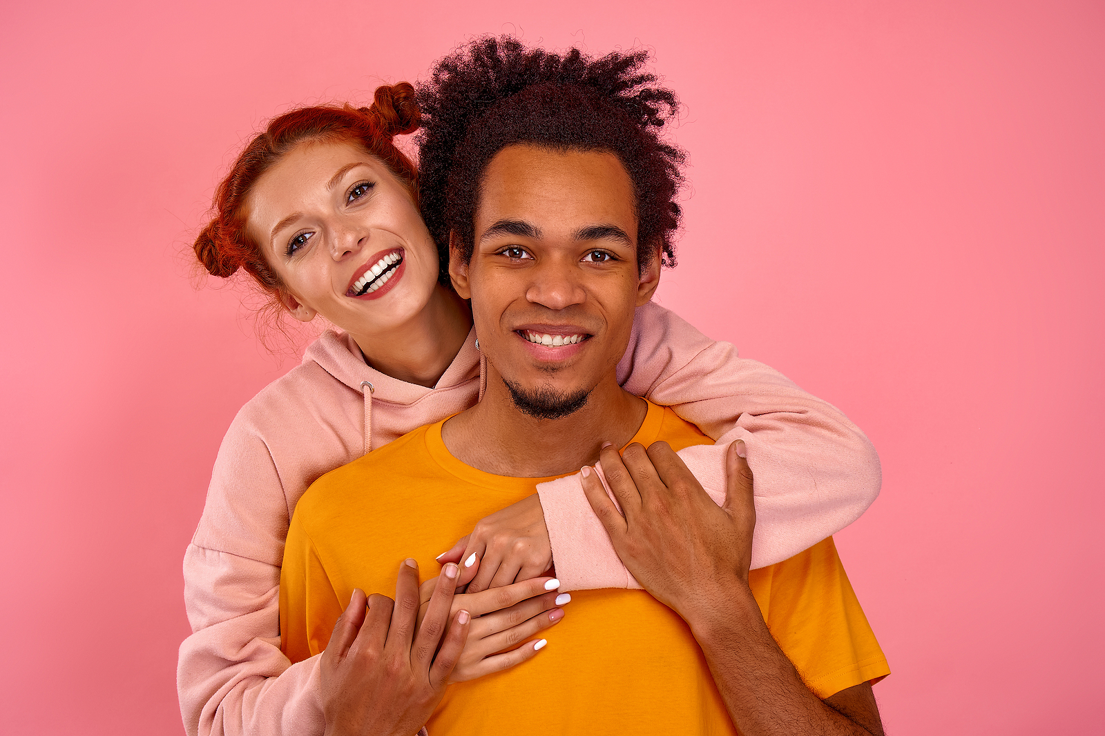 A woman stands behind her boyfriend hugging him and smiling while he holds her arms and smiles.