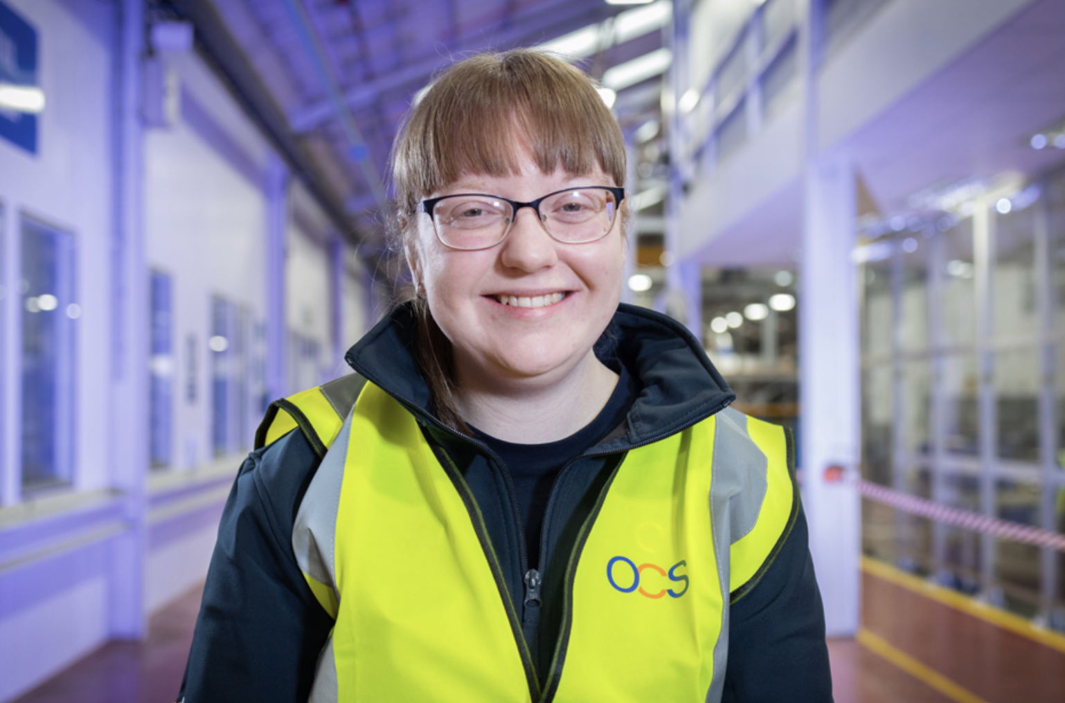 A smiling person wearing glasses and a yellow high-visibility vest with the OCS logo stands indoors in a brightly lit industrial or warehouse setting.