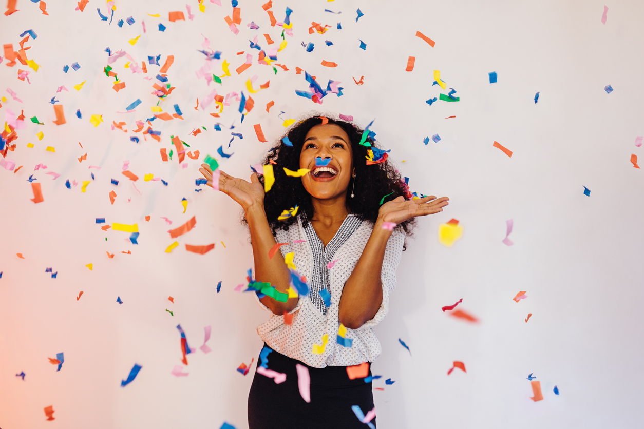 Image of a young attractive black woman with long hair throwing confetti into the air and smiling against a plain background.