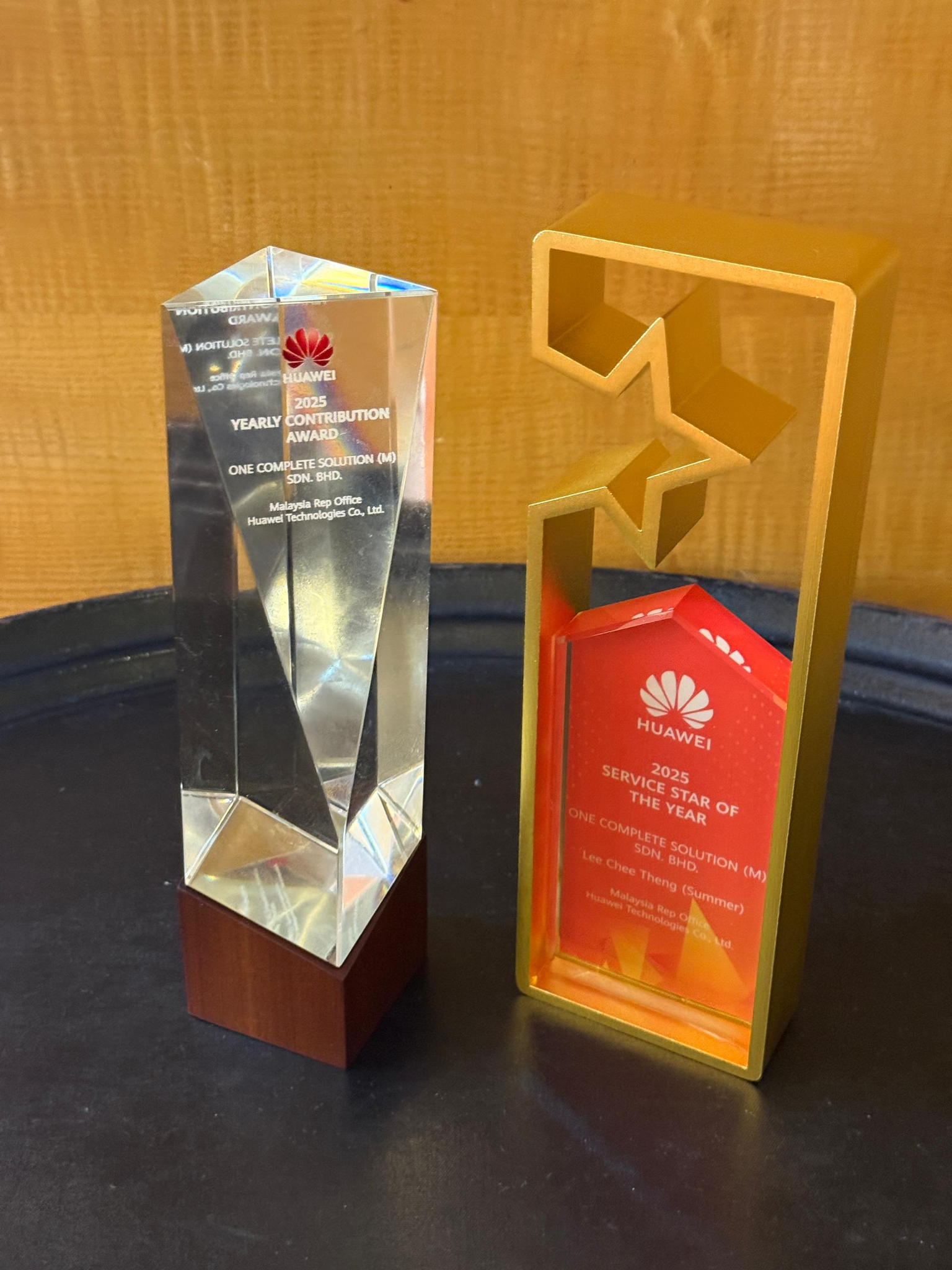 Two awards are displayed side by side on a wooden surface. The left is a clear, faceted glass trophy with a wooden base. The right is a red and gold geometric trophy labeled Huawei 2023 Service Star of the Year.