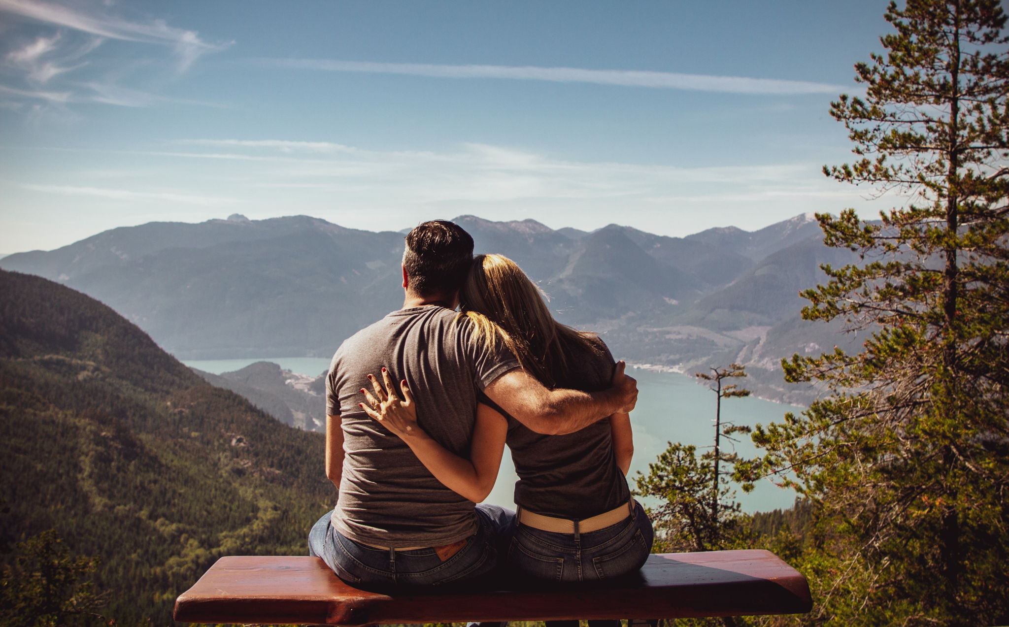 A man and a woman sit on a bench overlooking a beautiful scenery holding eachother.