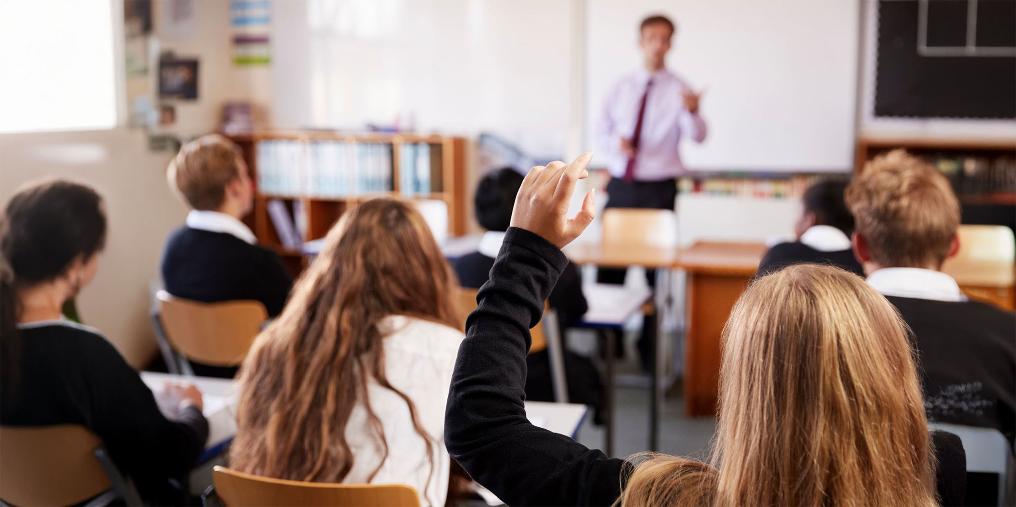 A student raises their hand in a classroom while others sit facing a teacher, who stands at the front near a whiteboard, leading a lesson. The students appear attentive and engaged.