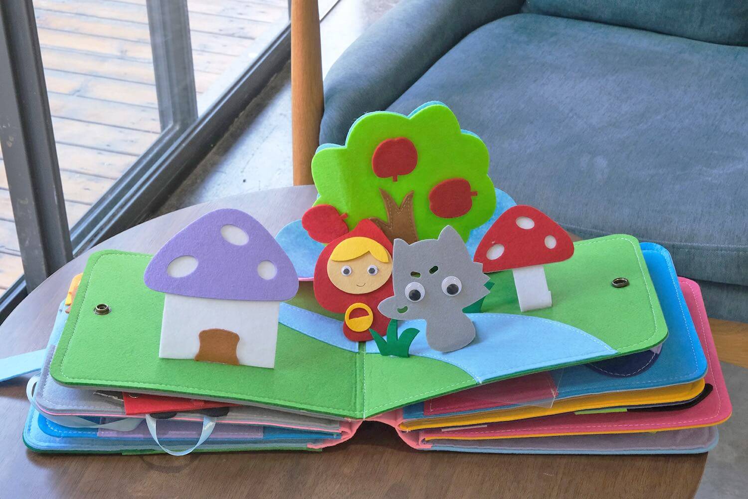 An opened page of a quiet book placed on a table showing a house, a tree, a wolf, and a Little Red Riding Hood.
