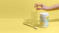 Woman's hand reaching out to stir a glass of ice water with a straw, next to a jar of Brightcore's Revive, a high quality, organic multi-collagen supplement in powder form, on a yellow background