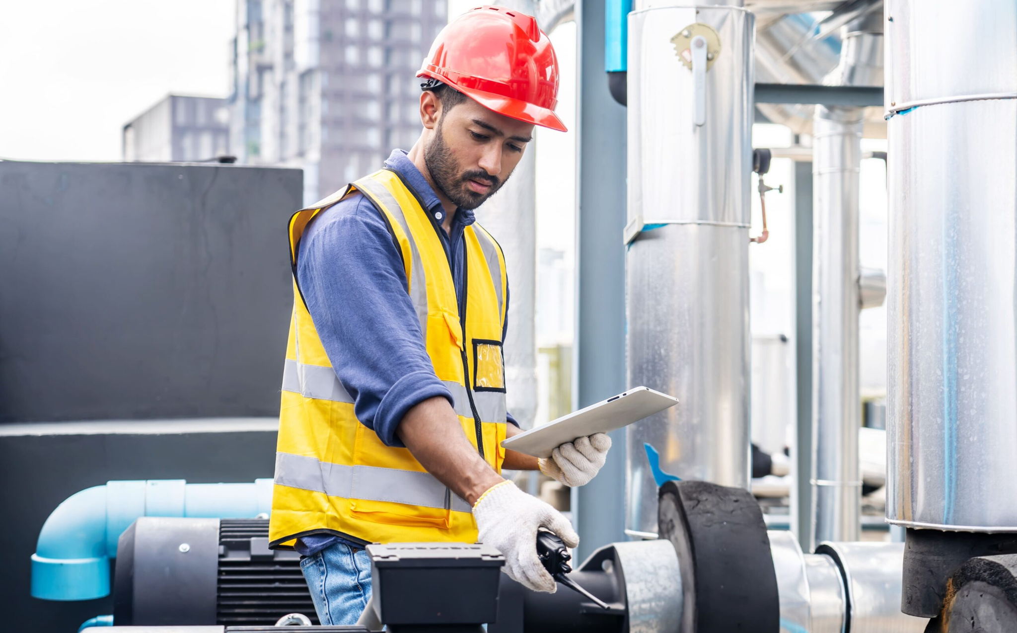 A male engineer wearing a red hard hat, yellow safety vest, and gloves inspects industrial equipment outdoors while holding a clipboard and using a handheld tool. Buildings and pipes are visible in the background.