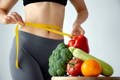 woman measuring her waist after improving her diet with a green powder supplement, vegetables on cutting board at front of photo