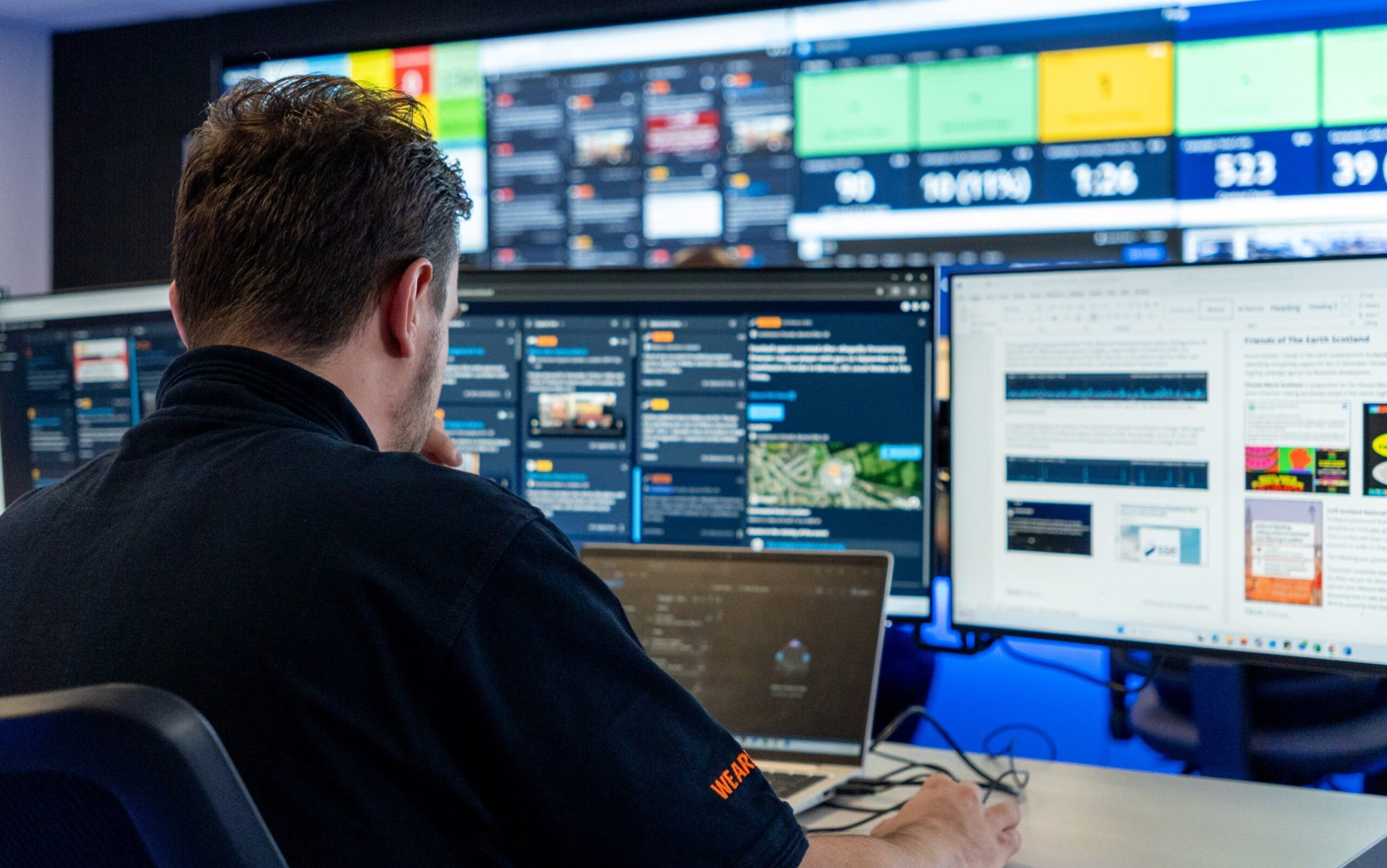 A person sits at a desk with multiple computer monitors displaying charts, graphs, and data, working in a control room with large screens showing dashboards and information in the background.