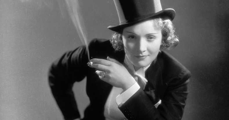 Image of Marlene Dietrich in her signature suit, and a cigarette in her hand, smirking at the camera.