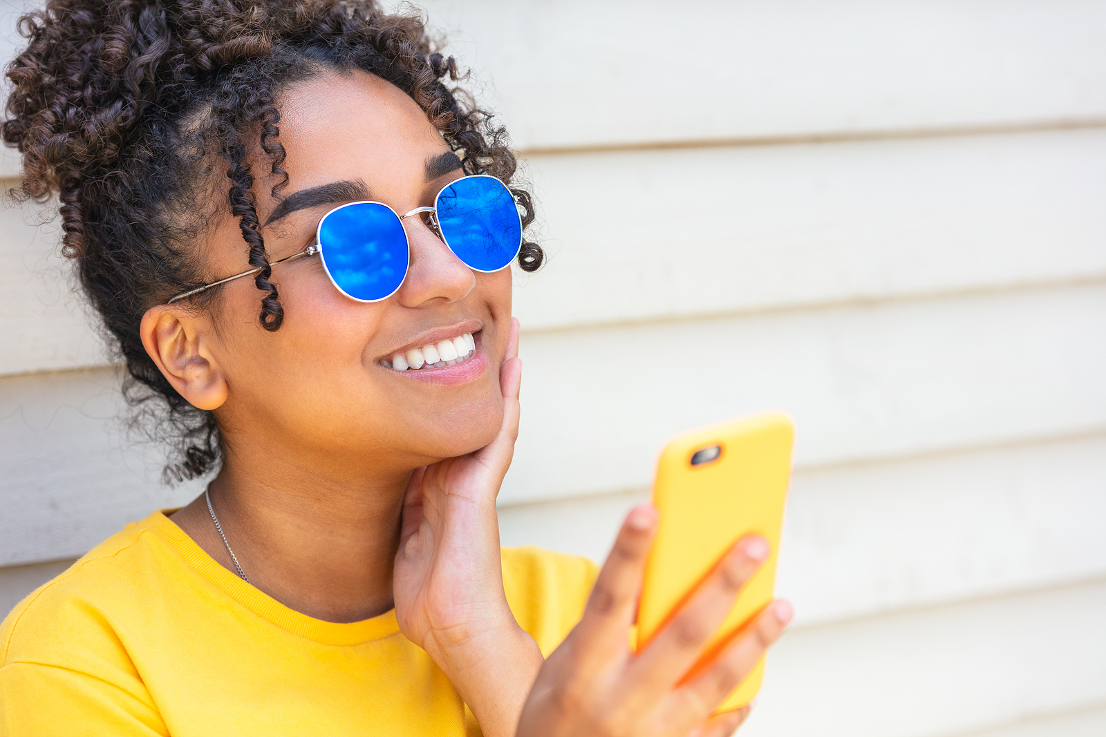 An attractive black woman with sunglasses hold her phone up and smiles for the camera.