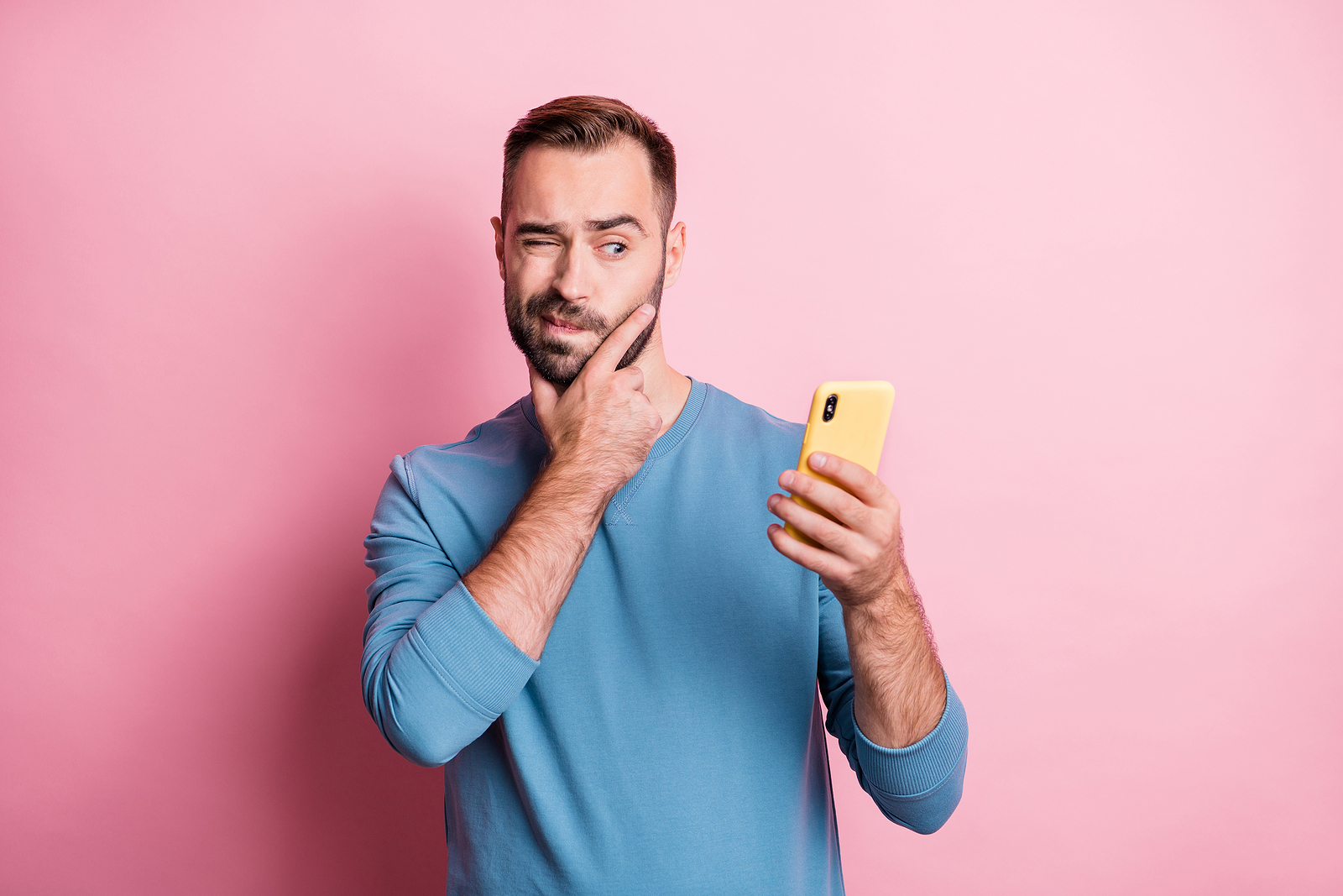 A man looks at his phone with one eye while contemplating, holding a hand to his chin against a pink background.