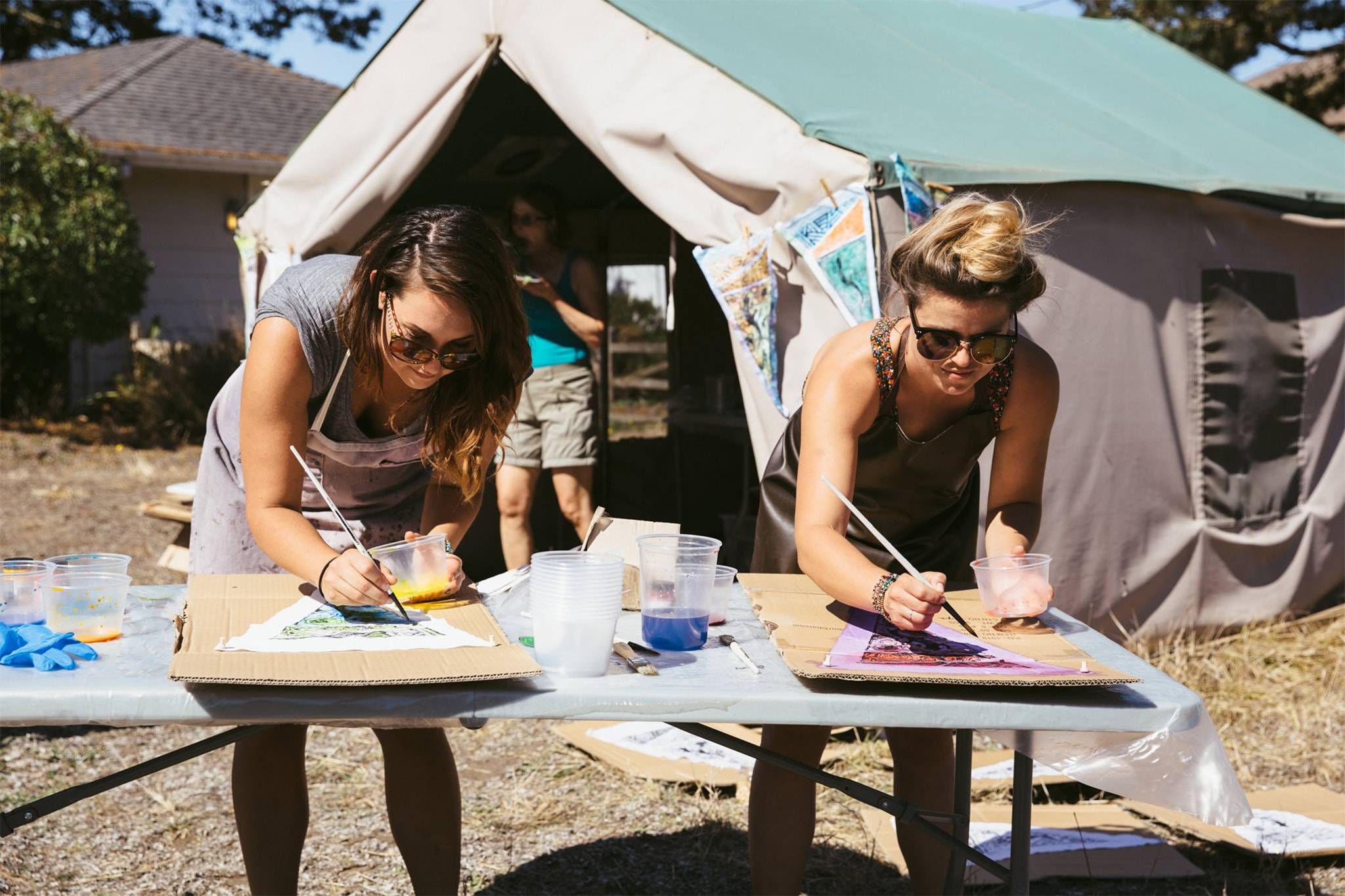 Two women painting on a box outside their tent