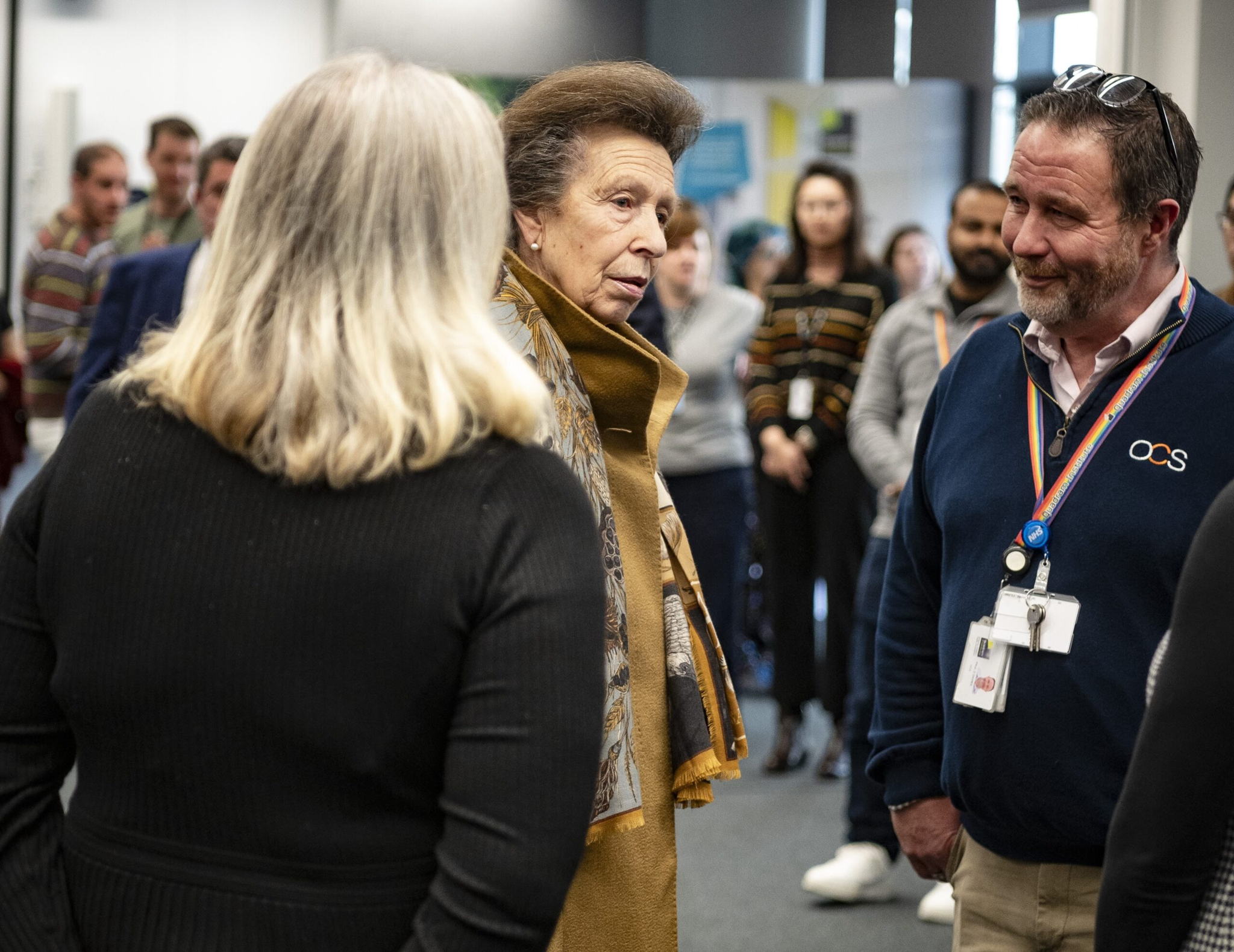 A group of people engaged in conversation indoors. One person in the center is wearing a brown coat and scarf. Others stand around, some with lanyards. The background shows blurred figures and interior elements.