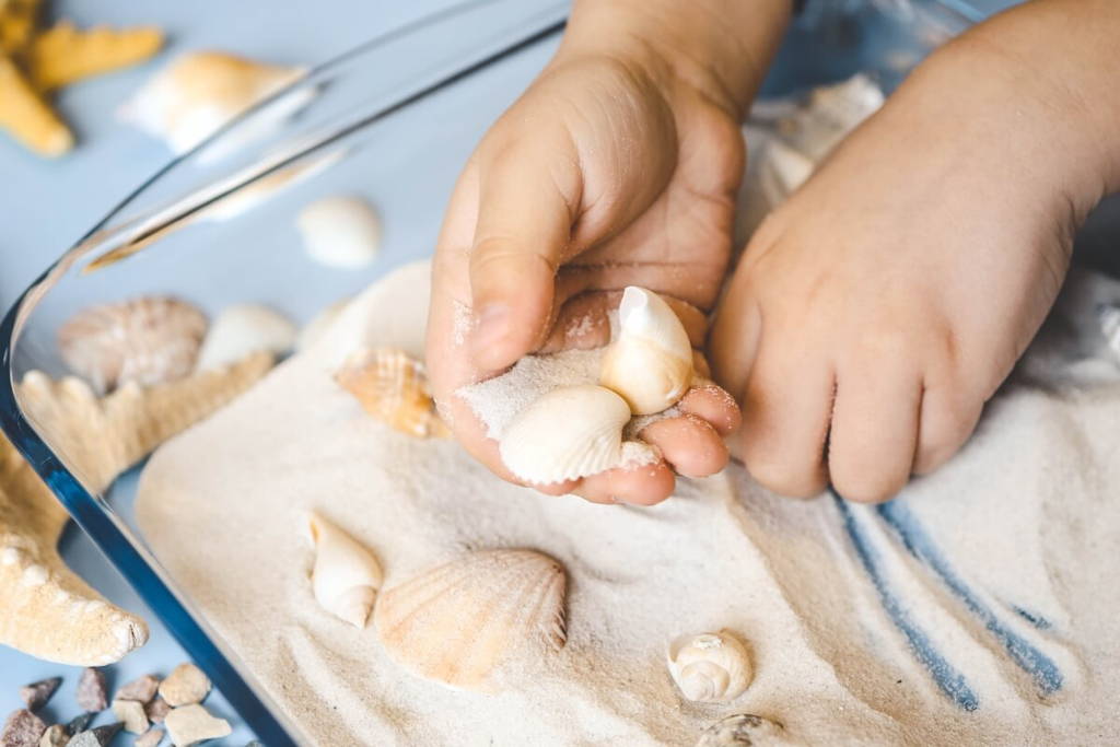 Child playing with sand and sea shelves that are placed in a glass sensory bin.