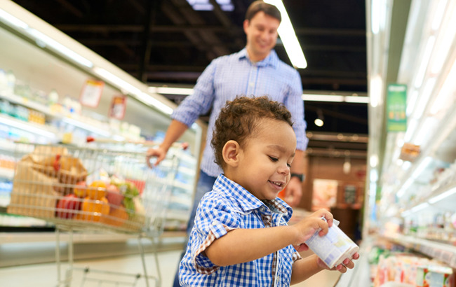 Little toddler boy picks up a pack of yogurt in the supermarket as his father looks on