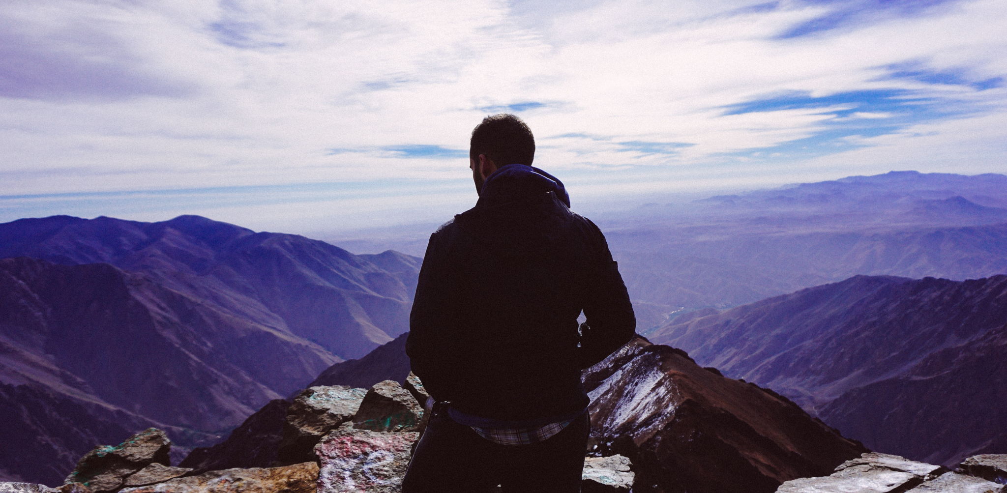 The silhouette of a man standing at the top of a hill overlooking the mountains.