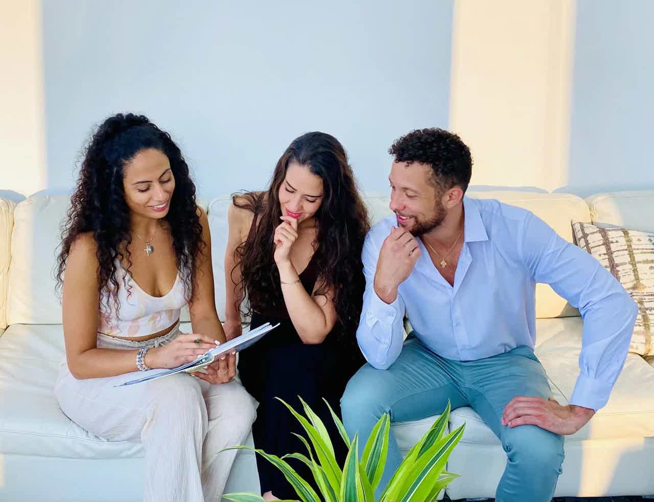 Three people sitting on a sofa, looking at a tablet together in a bright room.