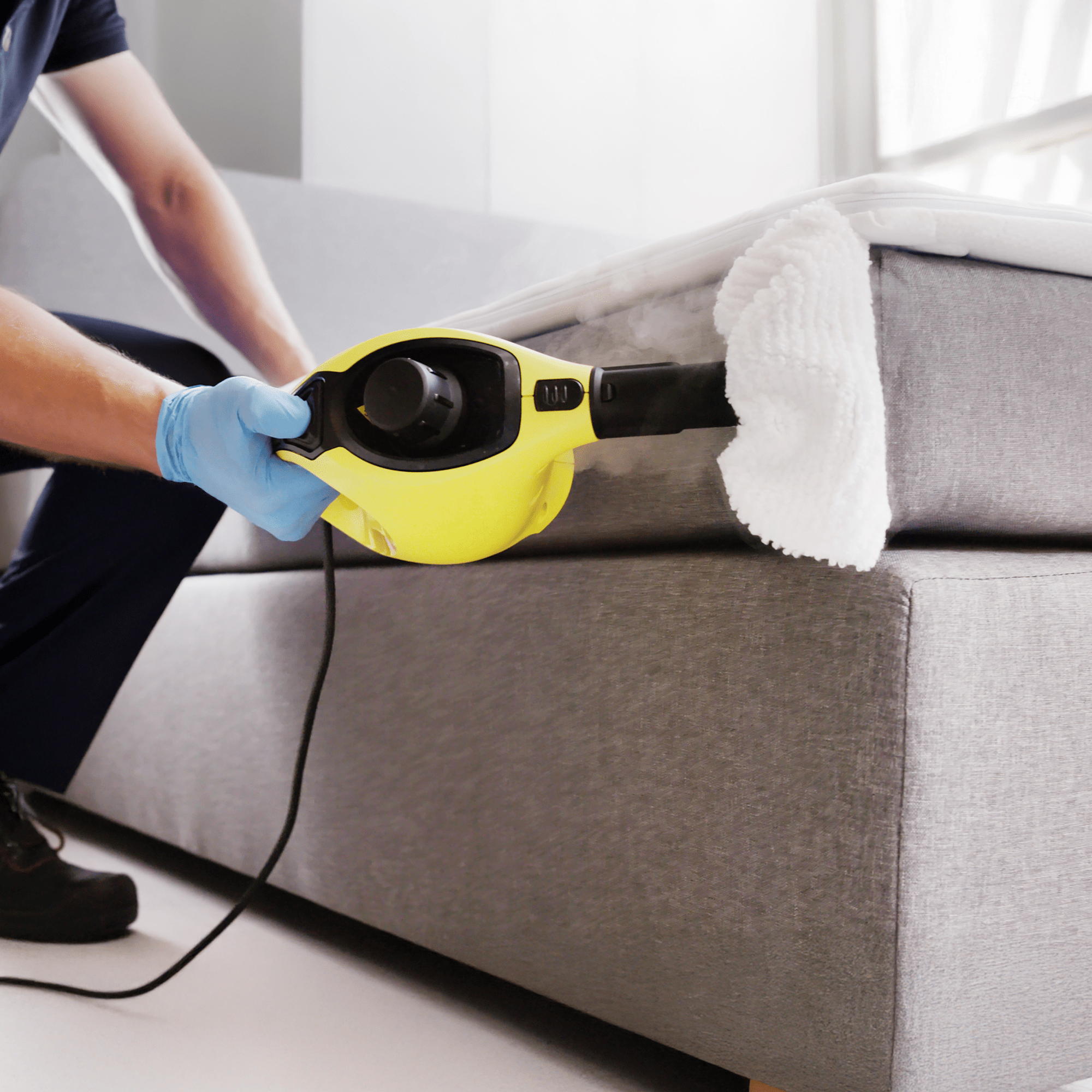 A person wearing a blue glove uses a handheld steam cleaner with a white cloth attachment to clean the side of a gray upholstered bed.