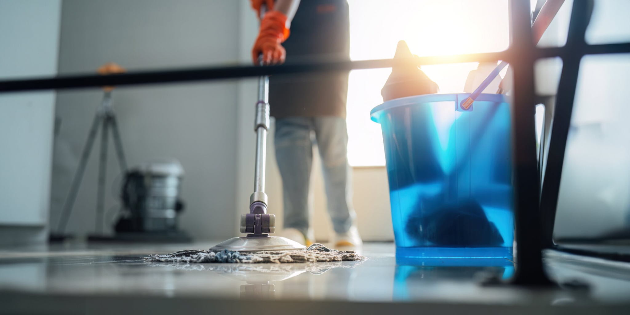 A person wearing orange gloves mops a shiny floor near a blue bucket filled with cleaning supplies, with sunlight streaming in from a window in the background.
