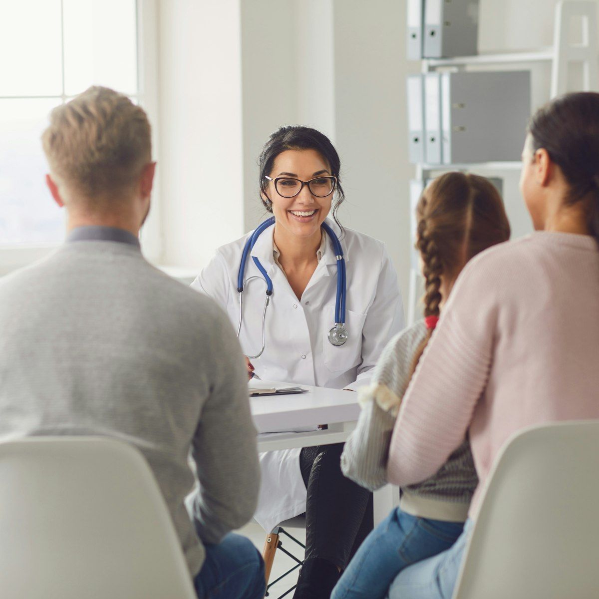 A smiling doctor with a stethoscope around her neck sits at a desk, talking to a family of four—two adults and two children—during a medical consultation in a bright office.